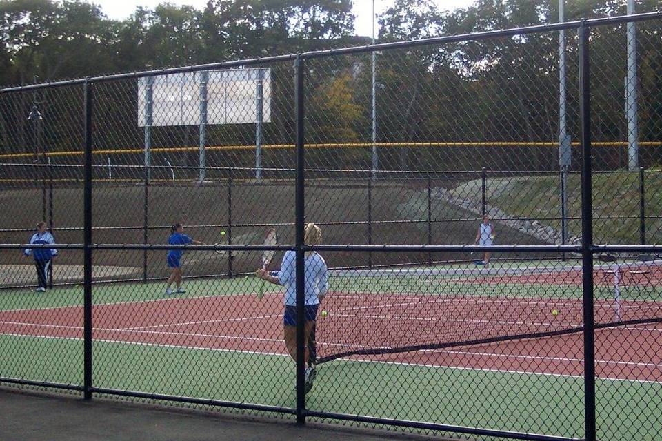 Several players are tennis in the tennis court enclosed by black PVC coated chain link fence.