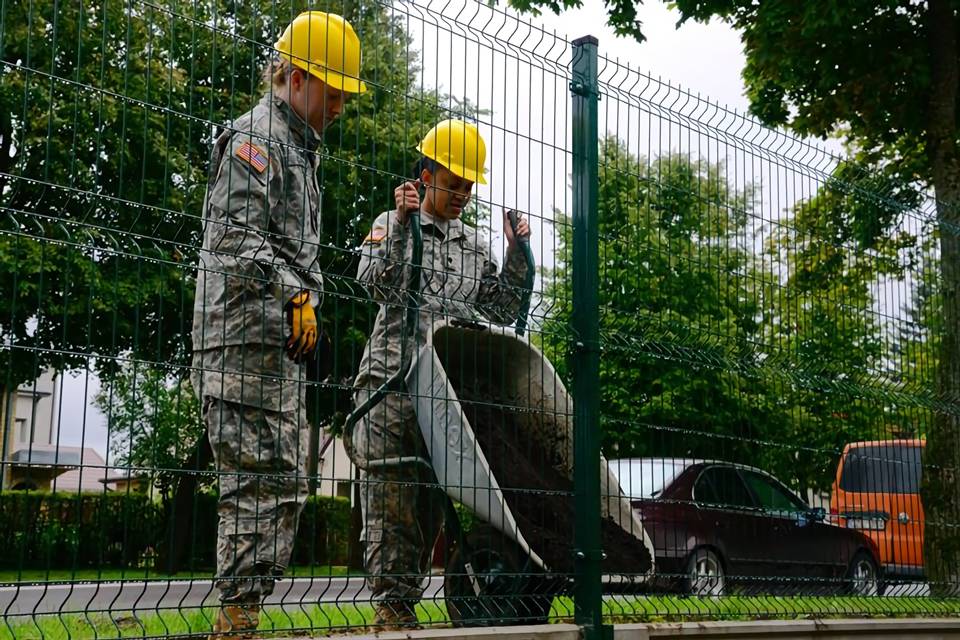 Two soldiers load concrete for curvy welded military fence construction.