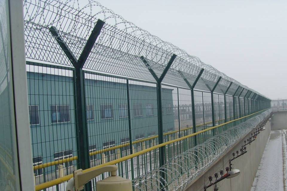 Curvy welded fence is installed on the wall and equipped with a concertina topping.